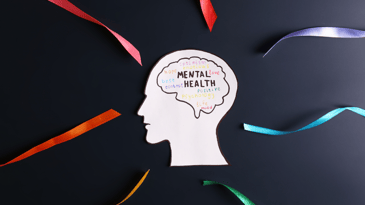 A paper cutout of a persons head with the brain drawn in the center with the words mental health written inside. Other words such as hope, positive, mood, love, and control are written in cursive in various bright colors.