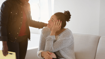 A patient looks up at their counselor who has their hand on their shoulder in a sign of support in a professional setting with natural light.