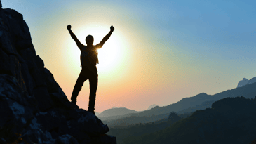 A person in silhouette with their arms up in a victorious gesture stands on the side of a mountain at sunset.