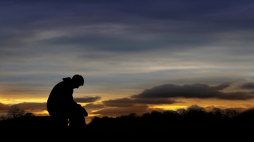 A person silhouetted by sunset sits on a rock with their head down and their hands hanging loosely in a sign of defeat. Clouds gather in the distance.