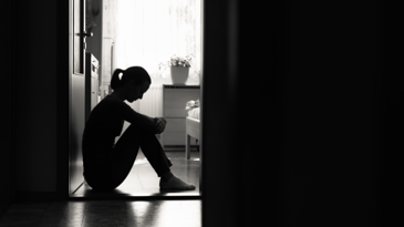 A person sitting on the ground with their back against an open door is silhouetted by the light coming through the window.