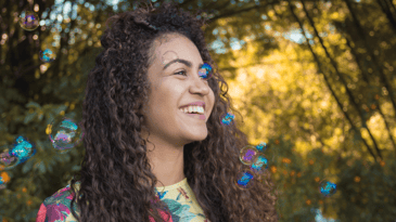 A person with brown curly hair smiles while standing in a natural setting with bubbles around them