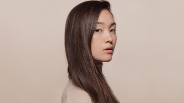 A person with brown hair and brown eyes looking sideways at the camera against a light pink background