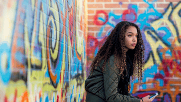 A person with dark curly hair holding a mobile device and sitting near a wall covered in graffiti