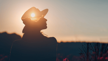 A silhouette of a person smiling off to the right in an outside setting wearing a wide-brimmed hat with the sun and clouds on it.