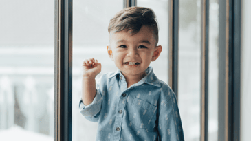 A small child with dark hair and eyes smiling with their hand up against a backdrop of a cityscape seen through a large window