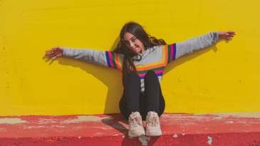A teenager holding their arms out against a bright yellow wall in the sun. They're wearing a rainbow sweater, black tights, and pink sneakers