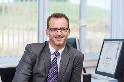 Friendly businessman wearing glasses sitting at his desk in the office looking at the camera with a smile