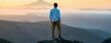 A person in a light blue shirt and tan pants looks out over a foggy, mountainous landscape at sunset. 