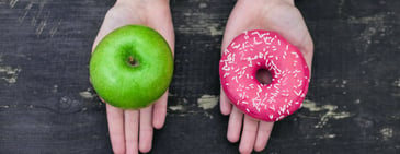 Two hands with a pink donut on one hand and a green apple in the other. 