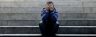 A person sitting on gray steps holding their head up with their hands in a pose indicative of sadness, anxiety, or depression. 