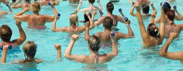 A group of people lifting light hand weights over their head during an swim exercise class in a bright outdoor swimming pool. 