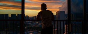 A person wearing a baseball cap looking out over a cityscape at sunset. 