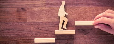 A wooden cutout of a human walking up step with a person's hand holding the next step in place. 
