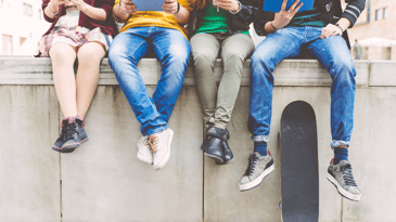 The legs of four teens reading and sitting on a wall. Each one wears distinctive sneakers and pants.