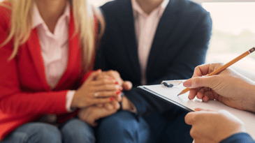 Two people hold hand in a blurred background while a persons hands holding a pencil and clipboard are clear in the forefront