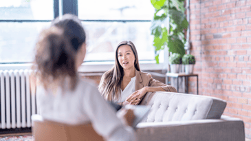 A person with long brown hair sitting on a sofa talking to a therapist in a room with natural light from large windows, brick walls, and plants.