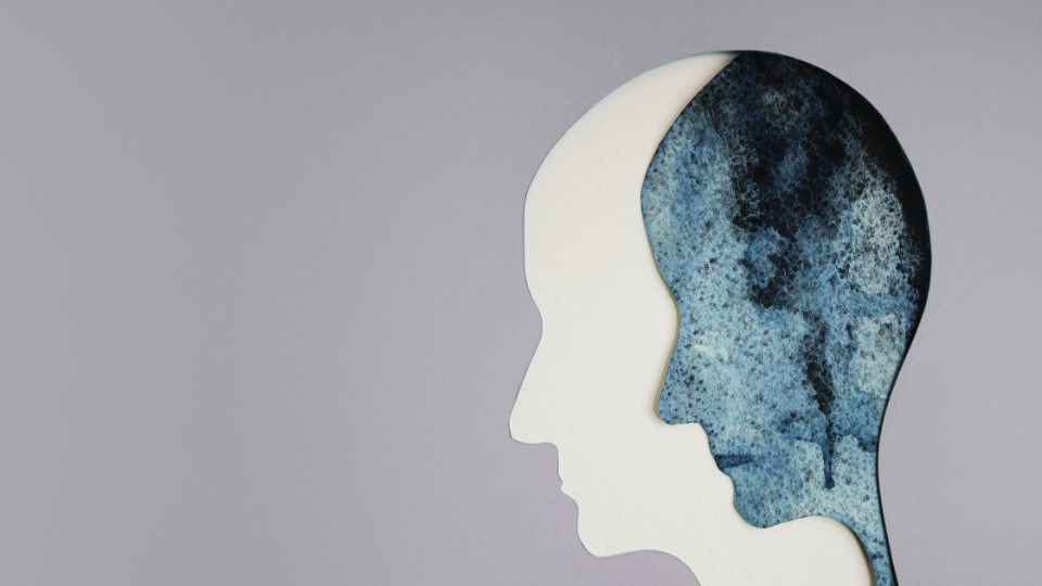 A gray cardboard cutout of a persons head against a white background with a twin cutout with deep blues and black watercolors halfway inside. An art piece that depicts depression or anxiety. 