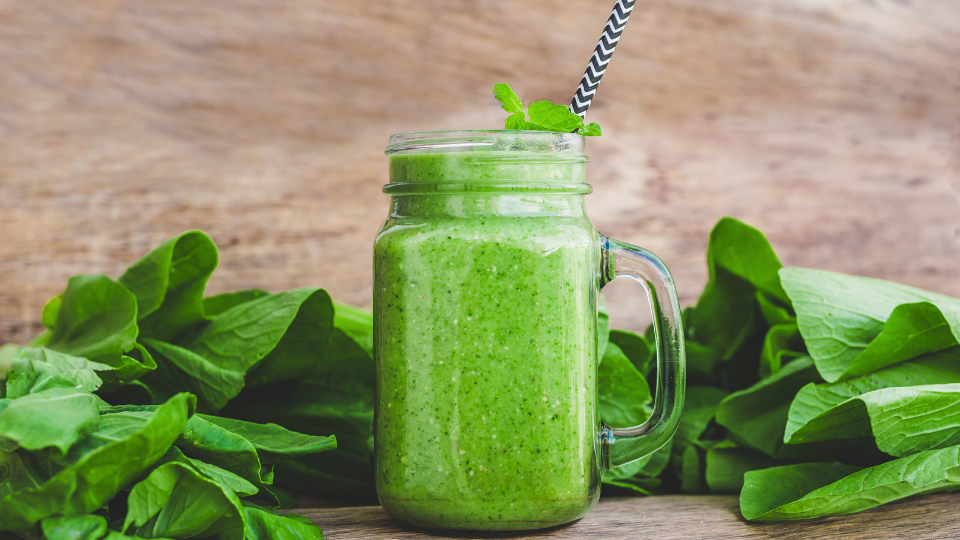 A green smoothie in a glass mug with a handle and a straw surrounded by kale leaves. 