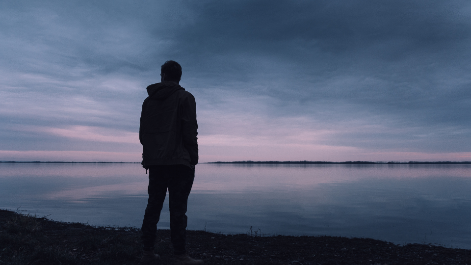 A person silhouetted against a purple and blue sunrise looks out across a lake.