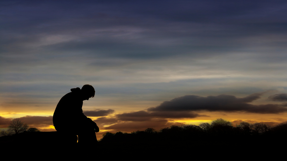 A person silhouetted by sunset sits on a rock with their head down and their hands hanging loosely in a sign of defeat. Clouds gather in the distance