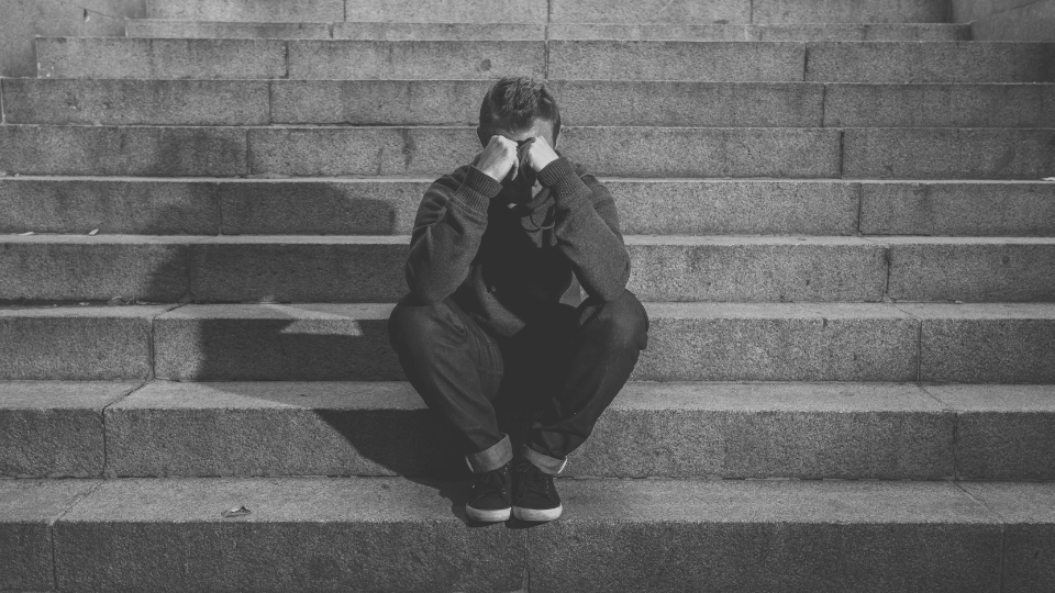 A person sits on gray steps with their hands balled in fists in front of their eyes in a sign of sadness.