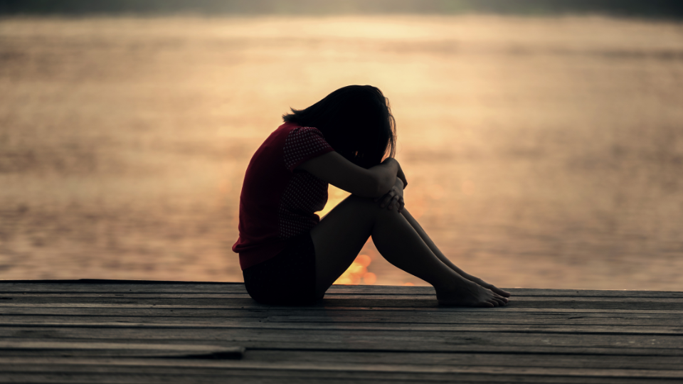 A person sitting on the dock of a lake crying with their head on their knees at sunset