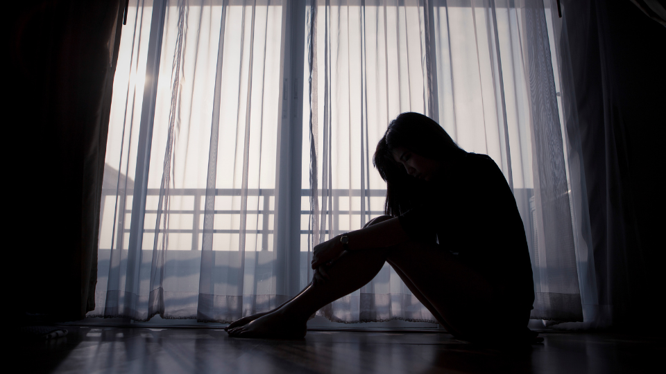 A person sitting on the floor next to a sliding glass door with transparent drapes hangs their head in sorrow