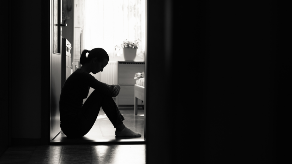 A person sitting on the ground with their back against an open door is silhouetted by the light coming through the window