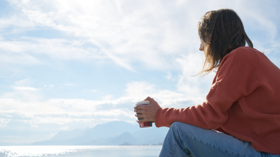 A person sitting outside looking over a landscape with water and white clouds with a cup of coffee in their hands
