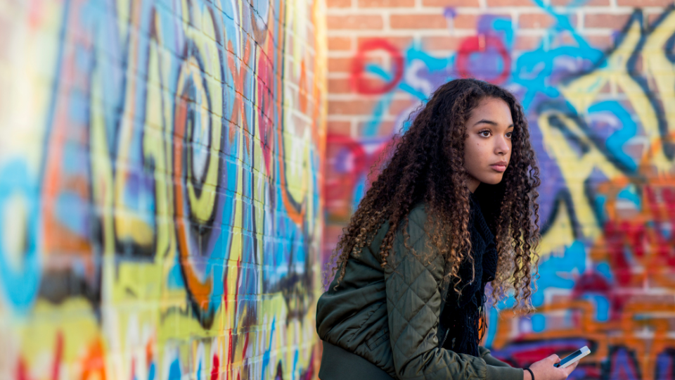 A person with dark curly hair holding a mobile device and sitting near a wall covered in graffiti.