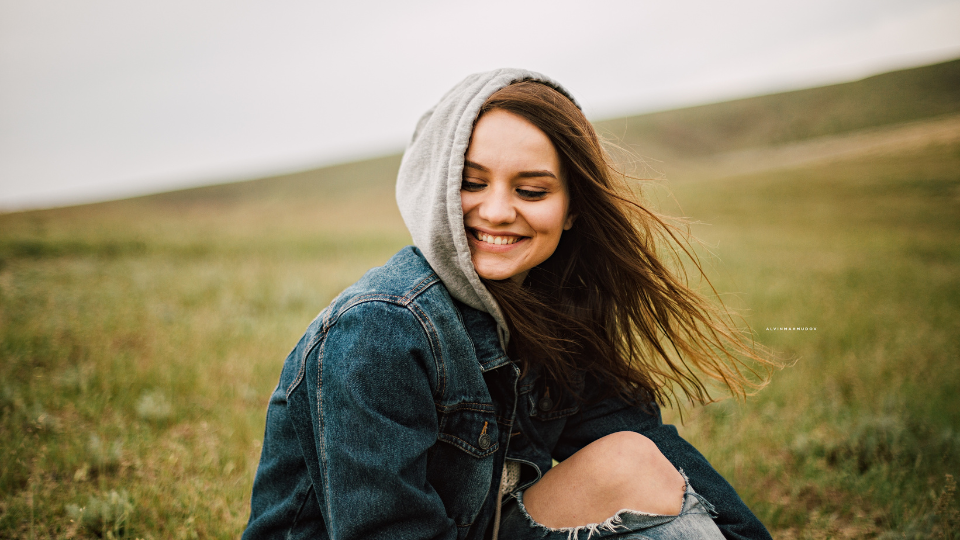 A smiling person sitting on a hillside wears a denim jacket over a gray hoodie.