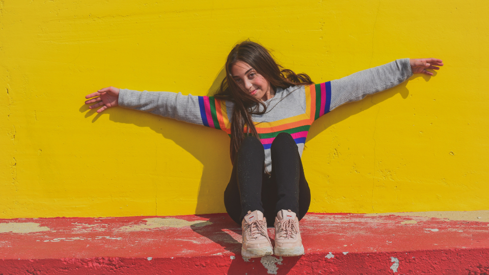 A teenager holding their arms out against a bright yellow wall in the sun. Theyre wearing a rainbow sweater, black tights, and pink sneakers.