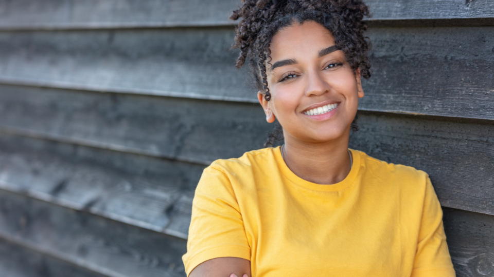 A teenager with curly black hair and dark eyes wearing a bright yellow t-shirt and smiling against a worn gray wooden wall