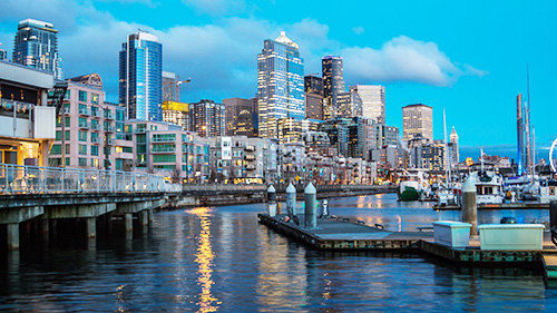 The Vancouver cityscape showing skyscrapers and lower apartment buildings against a clear blue sky as seen across a harbor.