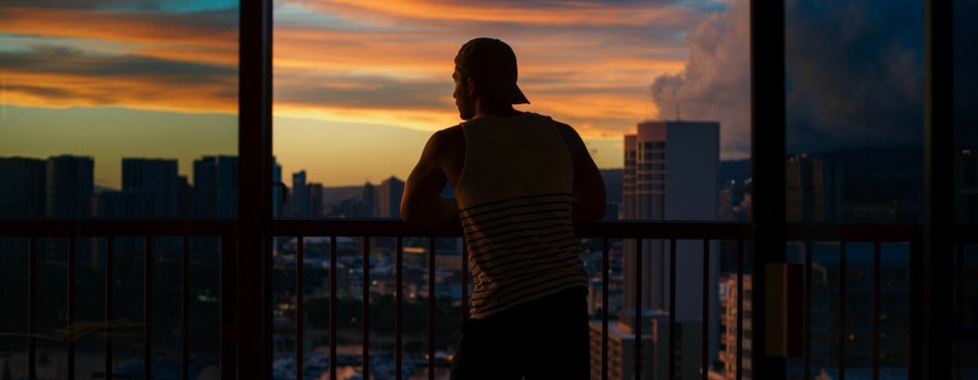The silhouette of a person wearing a baseball cap looking out over a city from an apartment balcony at sunset. 