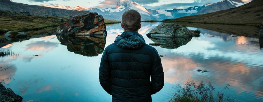 A person facing away from the camera toward a lake with snowcapped mountains. 