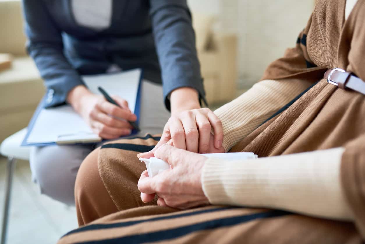 A person with a clipboard on their lap and a pen in one hand places one hand on another person's arm in a supportive gesture. The other person is holding a tissue in their hand as if they've been crying. 