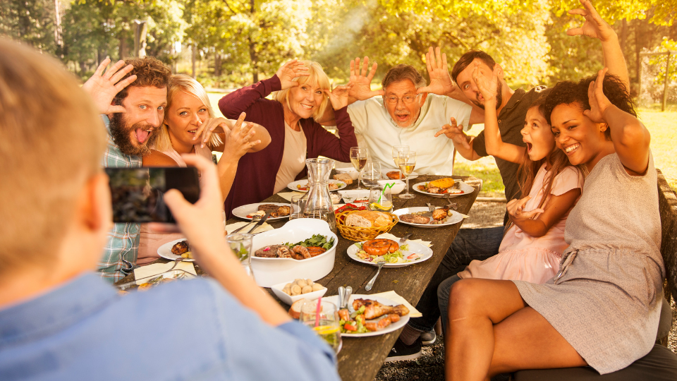 People sitting around a table for a holiday meal outside act silly while someone takes a family picture with a mobile phone. 