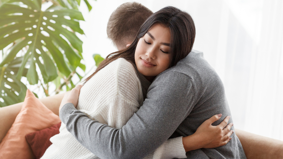 Two people hugging during a counseling session in a comforting setting with plants, pillows, and and natural light